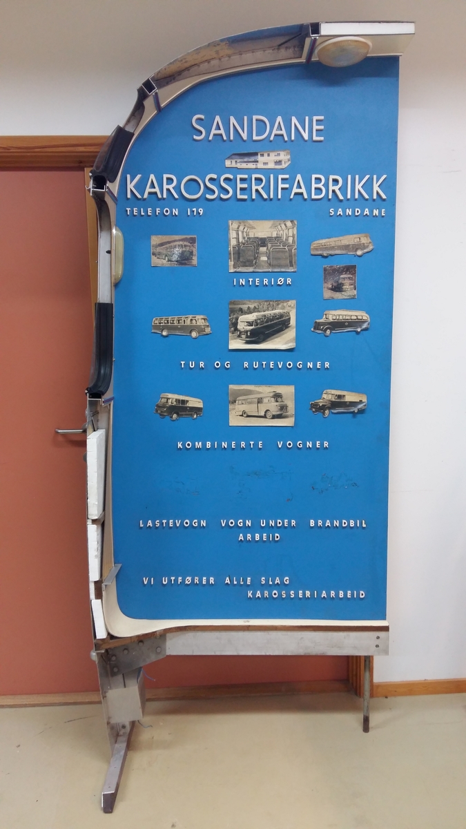 Reklameinstallasjon. Visningsmodell. Finèrplate påmontert snitt av karosseri på buss, med isoporisolasjon og gummlistar rundt vindauge, utside med pyntelistar, innside med kupèlys og vindauge med vindaugsisolasjon. Snittet går i ei boge på eine langsida og opp rundt langs øvste kanten, og er festa til ei finèrplate. Plata er blå på framsida, med pålimte bilete av bussar og andre bilar og tekst i gipsbokstavar. Installasjoen står på eit bein og ei støtte av metall.

 karosseriet på ein buss