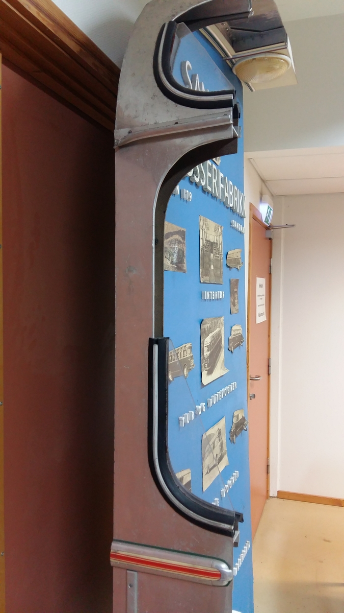 Reklameinstallasjon. Visningsmodell. Finèrplate påmontert snitt av karosseri på buss, med isoporisolasjon og gummlistar rundt vindauge, utside med pyntelistar, innside med kupèlys og vindauge med vindaugsisolasjon. Snittet går i ei boge på eine langsida og opp rundt langs øvste kanten, og er festa til ei finèrplate. Plata er blå på framsida, med pålimte bilete av bussar og andre bilar og tekst i gipsbokstavar. Installasjoen står på eit bein og ei støtte av metall.

 karosseriet på ein buss