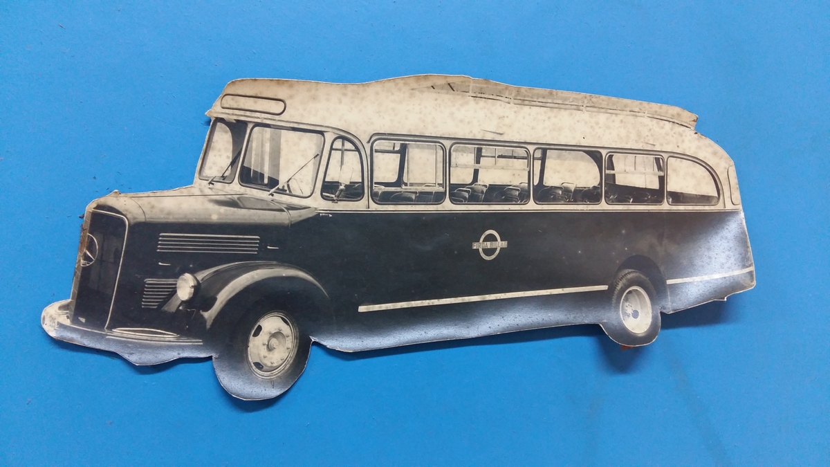 Reklameinstallasjon. Visningsmodell. Finèrplate påmontert snitt av karosseri på buss, med isoporisolasjon og gummlistar rundt vindauge, utside med pyntelistar, innside med kupèlys og vindauge med vindaugsisolasjon. Snittet går i ei boge på eine langsida og opp rundt langs øvste kanten, og er festa til ei finèrplate. Plata er blå på framsida, med pålimte bilete av bussar og andre bilar og tekst i gipsbokstavar. Installasjoen står på eit bein og ei støtte av metall.

 karosseriet på ein buss