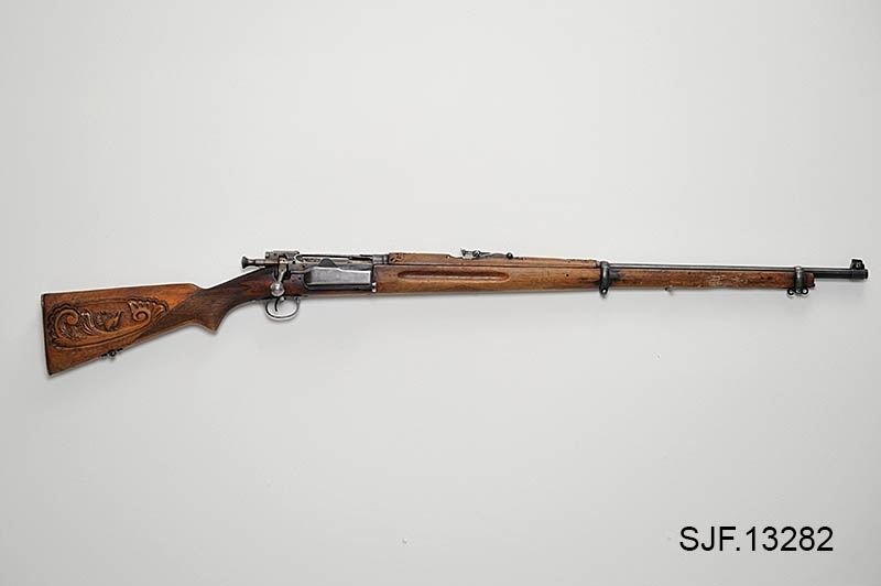 Krag-Jørgensen rifle med kaliber 6, 5 x 55. Geværet har pistolgrep. Magasinkapasitet på 5 patroner. Kolben er brunbeiset / oljet. Skjeftet er dekorert med forseggjorte utskjæringer.  Kolbekappen av stål har innfelt et lokk. Om man åpner lokkket kommer man til et "oppbevaringsrom" i kolben. Treverket som omslutter undersiden av geværløpet har noen sprekkdannelser.  Geværet er utstyrt med høydejusterbart skur og sidejusterbart korn. 