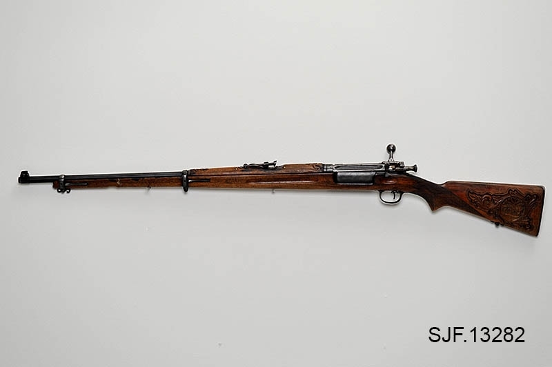 Krag-Jørgensen rifle med kaliber 6, 5 x 55. Geværet har pistolgrep. Magasinkapasitet på 5 patroner. Kolben er brunbeiset / oljet. Skjeftet er dekorert med forseggjorte utskjæringer.  Kolbekappen av stål har innfelt et lokk. Om man åpner lokkket kommer man til et "oppbevaringsrom" i kolben. Treverket som omslutter undersiden av geværløpet har noen sprekkdannelser.  Geværet er utstyrt med høydejusterbart skur og sidejusterbart korn. 