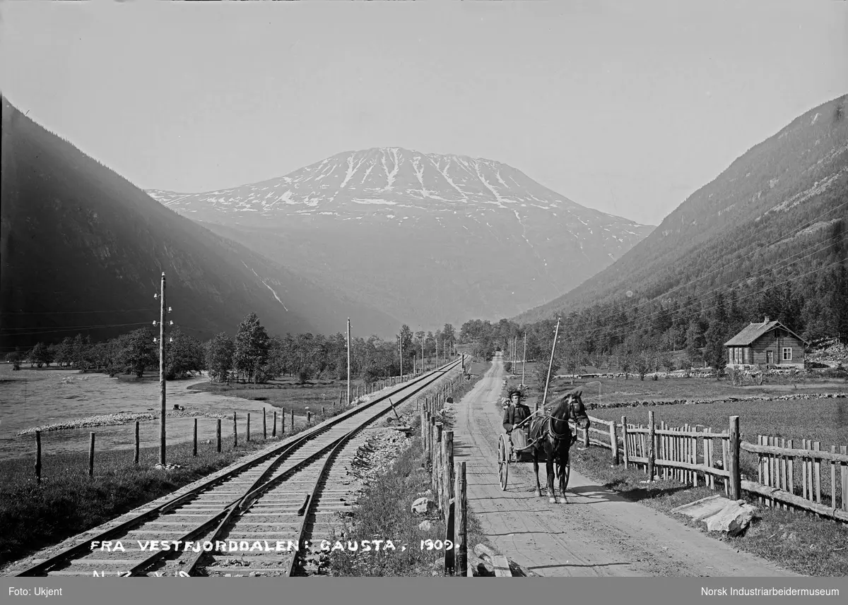 Hest med vogn og kusk på vei ved jernbanespor ved Bjørtuft. Gaustatoppen i bakgrunnen. Laftet bolig på høyre side.