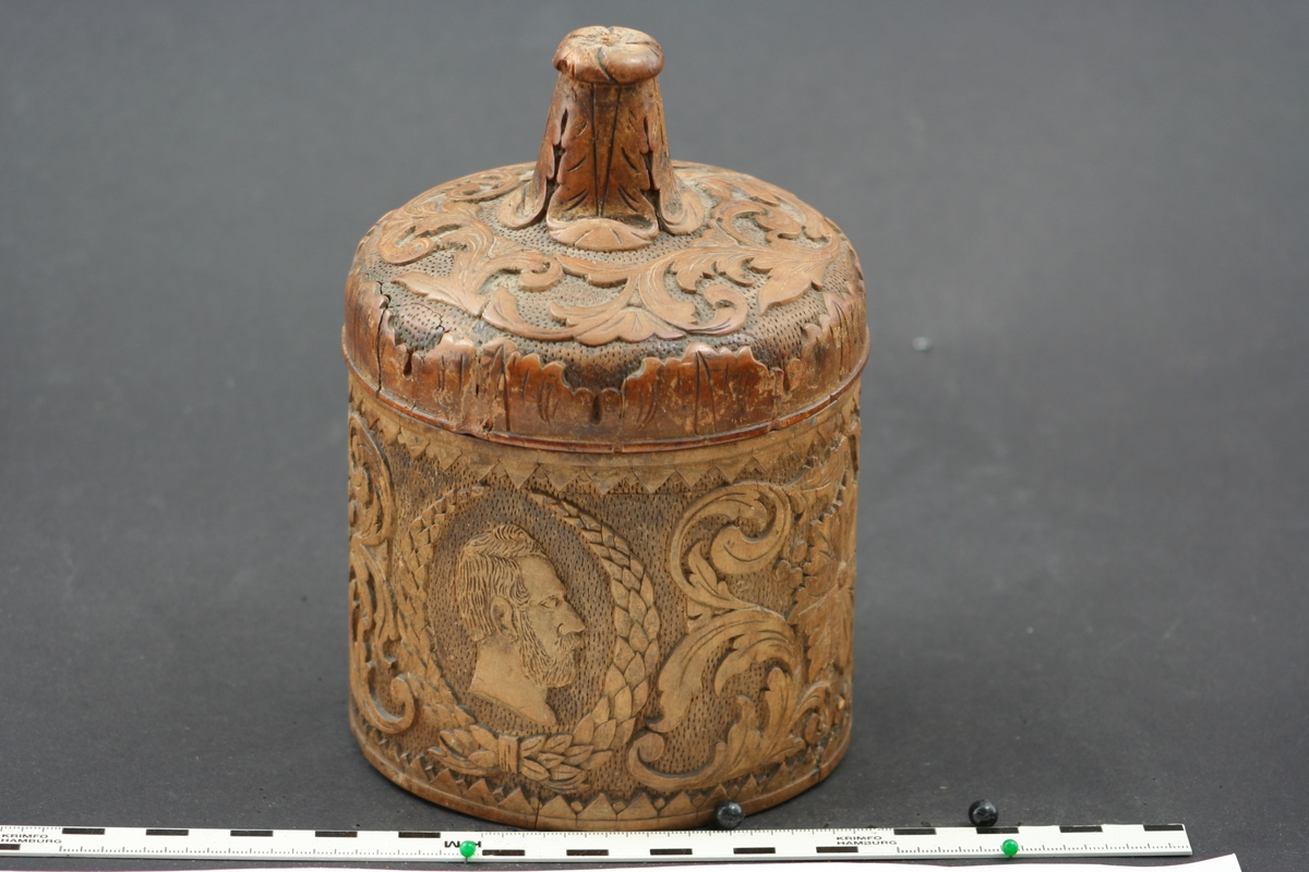 Tobaksburk av svarvad björk. Rund med skuren bladdekor, vapensköld, och porträtt av Karl XV inramad av en lagerkrans. Svagt välvt lock med hög knopp.  Material; Björk  Inköpt 1971 i antikaffär Gröna Terrinen(?) i Gamla Stan, för kr. 900 ;-