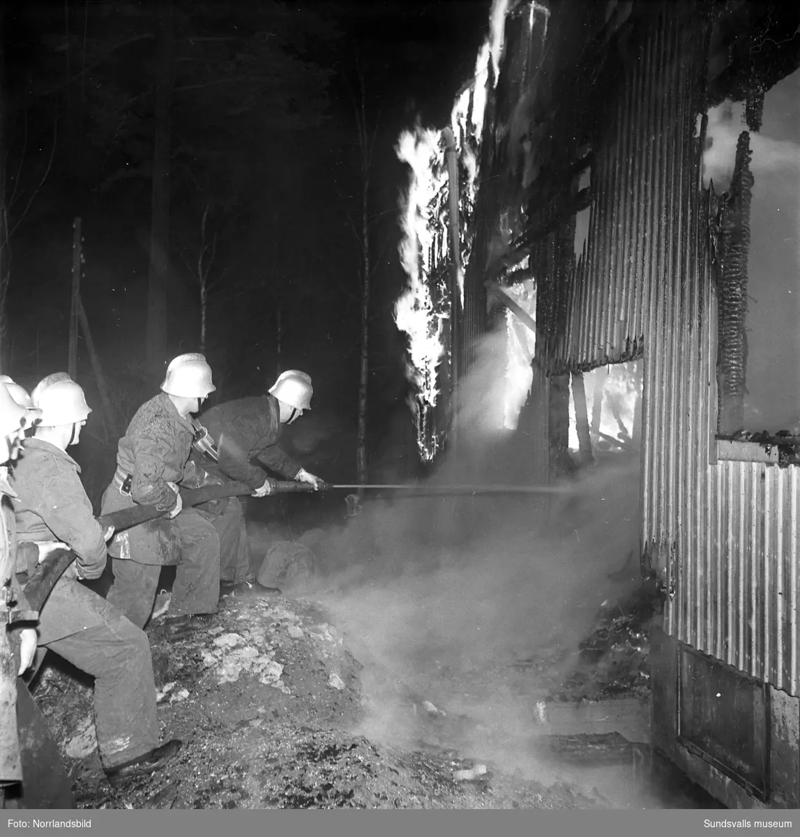 En fyrfamiljsfastighet invid Fagerviksfabriken är övertänd och brandmän från Fagervik och Vivsta sliter med släckningsarbetet. En stor samling åskådare har samlats. Huset höll på att renoveras för att ge husrum åt fabrikens tjänstemän.