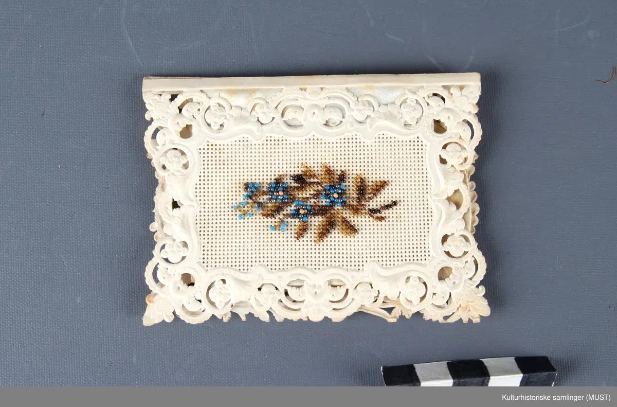 Nålemappe av perforert papp. Permene av hvit tynn presset pappkartong. På fremsiden initialene L.W. i perlebroderi. På baksiden et blomstermotiv i samme teknikk. Innvendig to "blader" av tynn filt, til nåler.