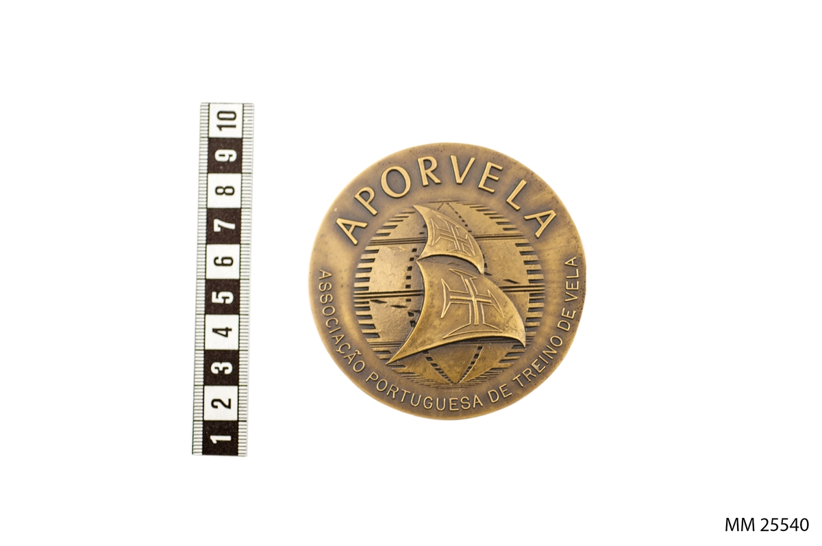 Rund plakett i metall med texten "Lisbon Sail - 1982" ovanför en relief av en borg och en stor rullande våg. Tillverkarsignaturen M. Barroco 82 står i nederkanten av denna våg. På andra sidan finns texten "APORVELA" och "ASSOCIACÂO PORTUGUESA DE TREINO DE VELA" runt en bild av segel med kors på.