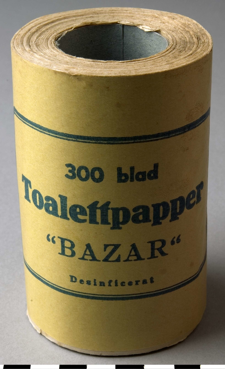 Toalettpappersrulle, 7 st, A-G.
Papprulle i mitten, omvirad med toalettpapper. Ytterst ett pappersomslag med texten: 300 blad. Toalettpapper, "BAZAR", Desinficerat. Omslaget är gult och texten i svart inom en svart ram.