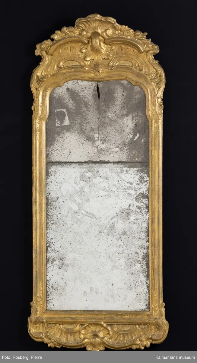 KLM 14346. Spegel, väggspegel. Förgylld ram av trä med snidade rokokoornament. Spegelglaset i två delar, facetterat, det övre spräckt. På baksidan Kalmar hallstämpel (ej den vanliga) samt otydlig signatur, troligen: Jonas Löw Kalmar. Rokoko.