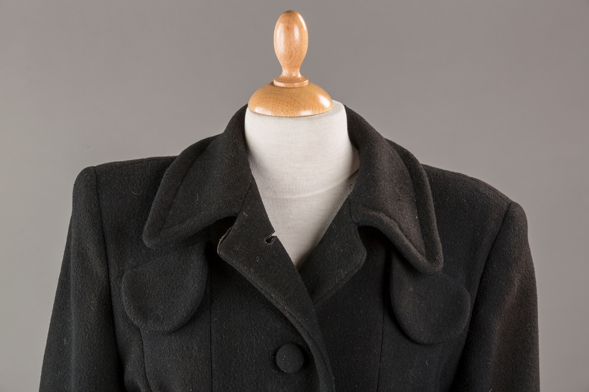 Innsvinget liv, pynteklaffer ved brystet foran. 
Stikklommer foran.
Skulderputer
Fire trukne knapper
Avrundet krage