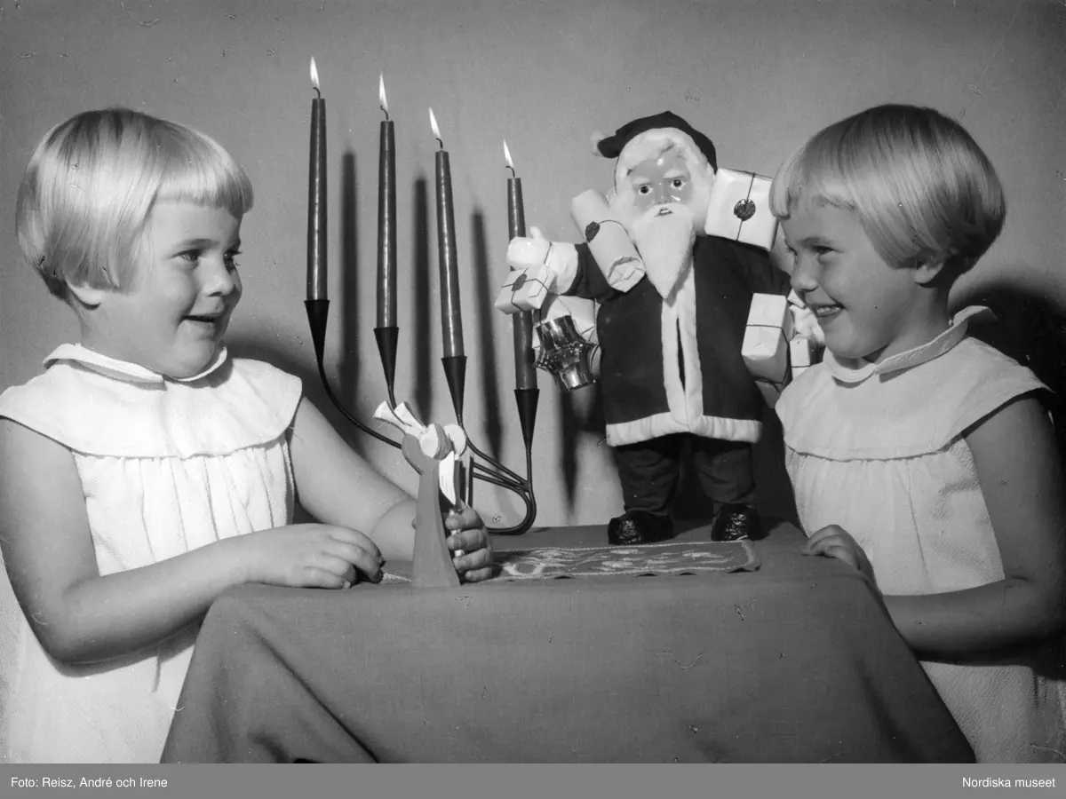 Två flickor i ljusa klänningar vid bord med adventsljusstake och tomtefigur