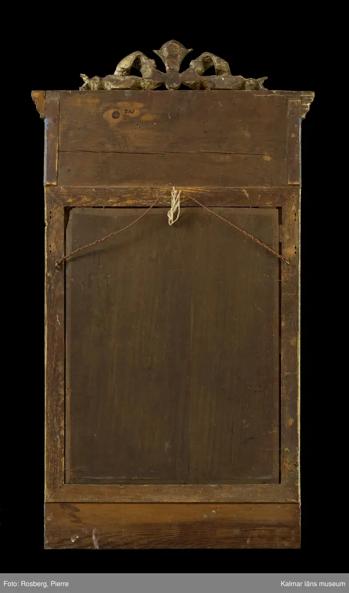KLM 45592. Spegel, väggspegel. Utförd i sengustaviansk stil. Skuren ornamentik i trä och i gjutet pastellage, förgylld med slagmetall. Ovanför glaset en rund medaljong med relief. Fasetterat originalglas. Träskuret krön i form av en avtagbar rosett.
