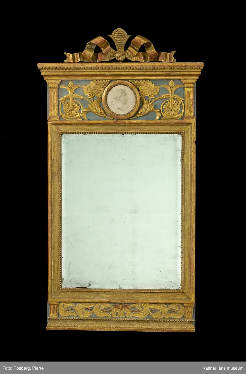 KLM 45592. Spegel, väggspegel. Utförd i sengustaviansk stil. Skuren ornamentik i trä och i gjutet pastellage, förgylld med slagmetall. Ovanför glaset en rund medaljong med relief. Fasetterat originalglas. Träskuret krön i form av en avtagbar rosett.