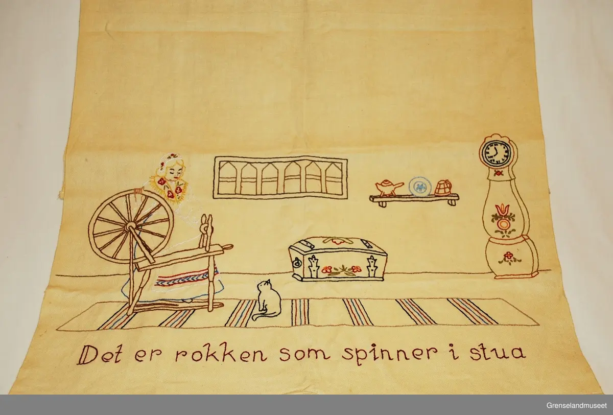 Brodert på gult bomullsstoff. Interiør med kvinne i bunadlignende drakt som spinner på rokk, kiste, gulvur, fillerrye og katt. Kontursting og tubgesting. "Det er rokken som spinner i stua"