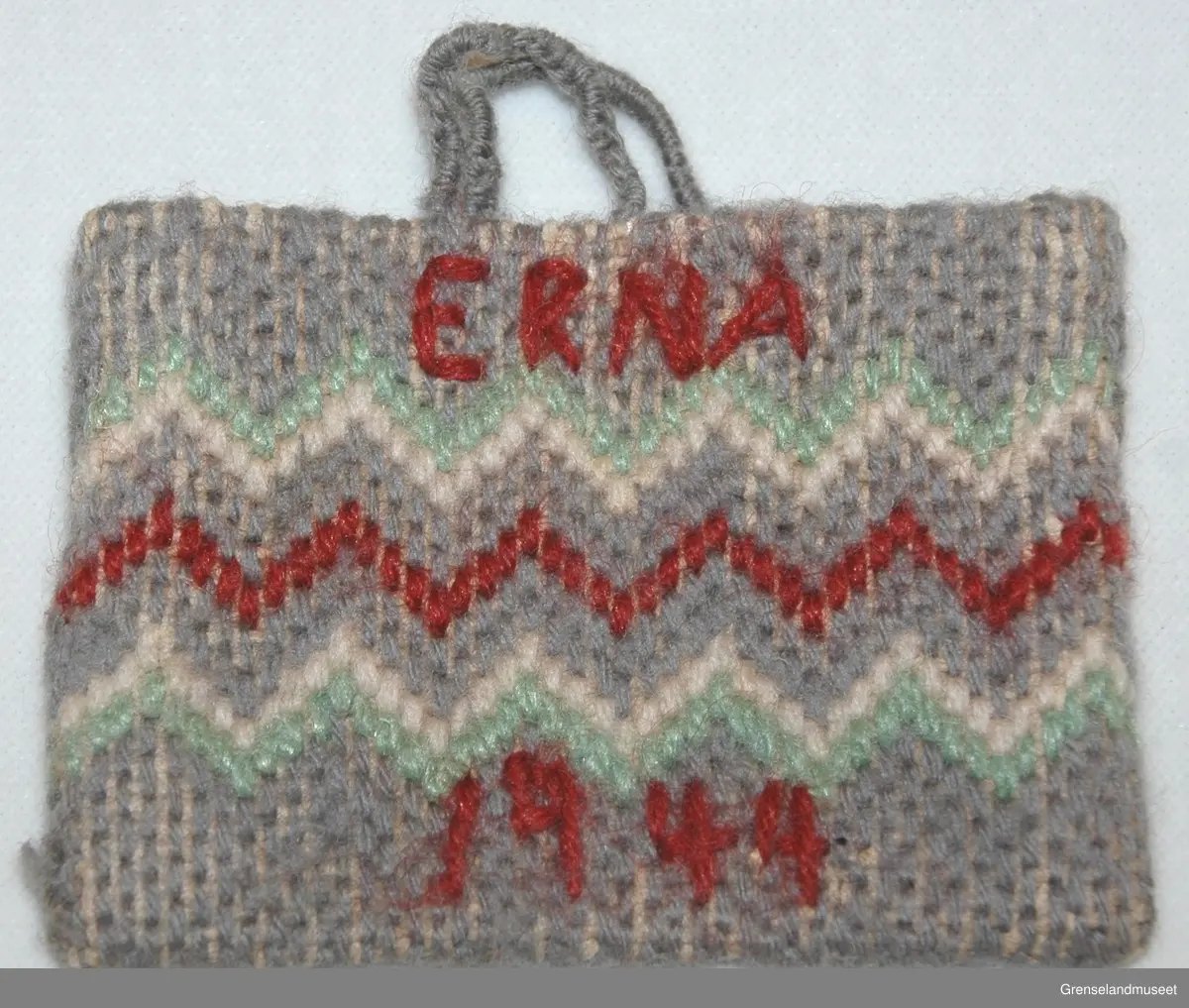 Brodert på stramei. Grå med hvite og rød sikksakkbord.
På den ene siden brodert: ERNA og 1944. På den andre siden: LUBECK