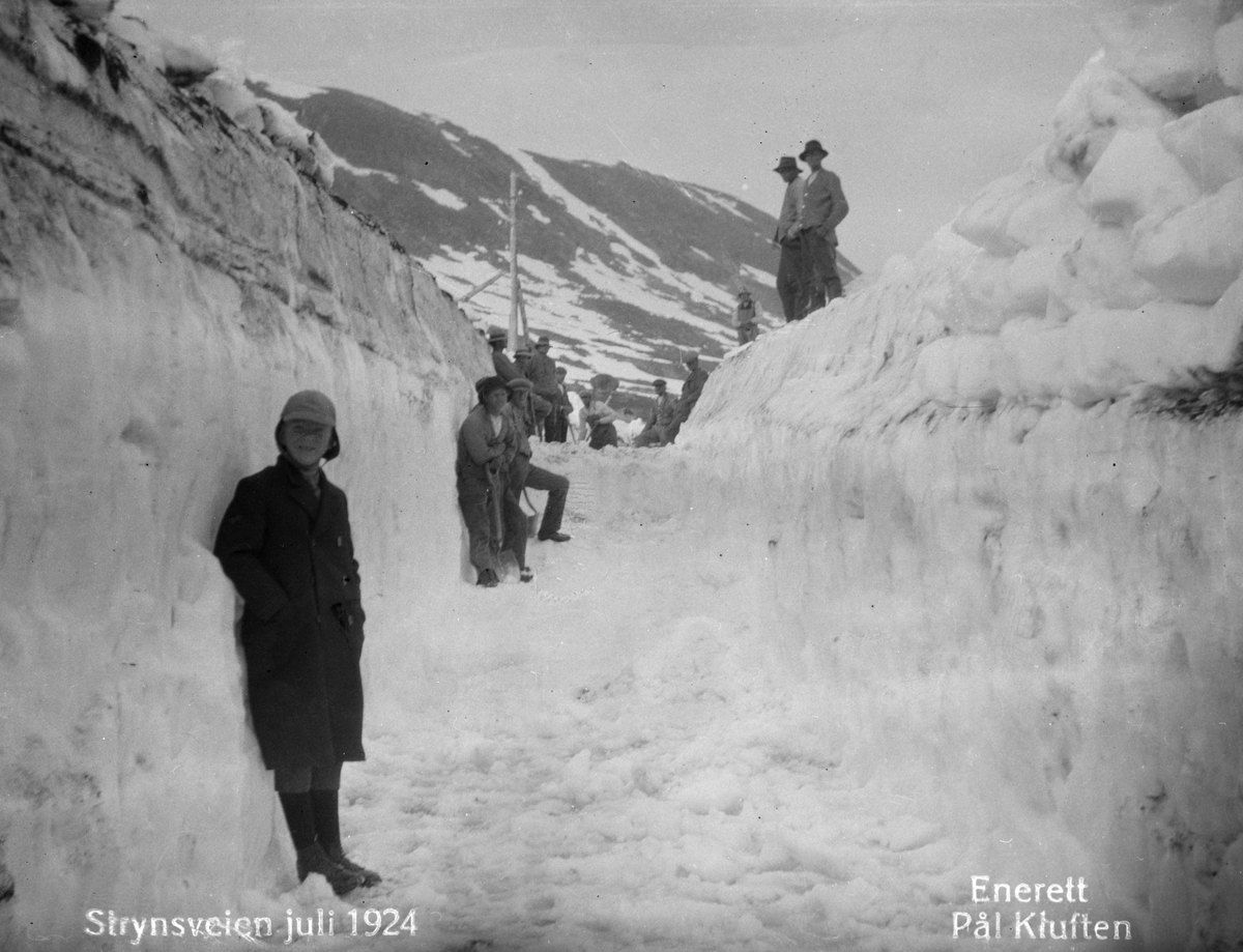 Snørydding av veg på Strynefjellet juli 1924. En gutt og to menn står nede vpå vegen med snøkant på godt over mannshøyde, mens flere mannfolk med spade står lenger bak.