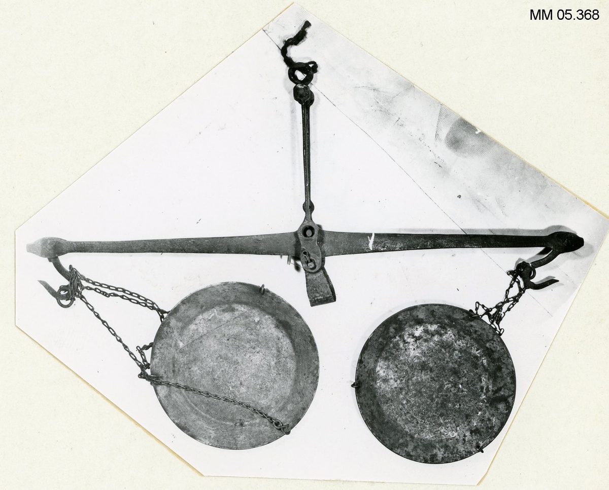 Vågbalans smidd av järn, försedd med vågskålar av bleckplåt, upphängda i kedja av mässing. Vågskålarna runda. Två stycken försedda med tre stycken kedjor av järn för upphängning.