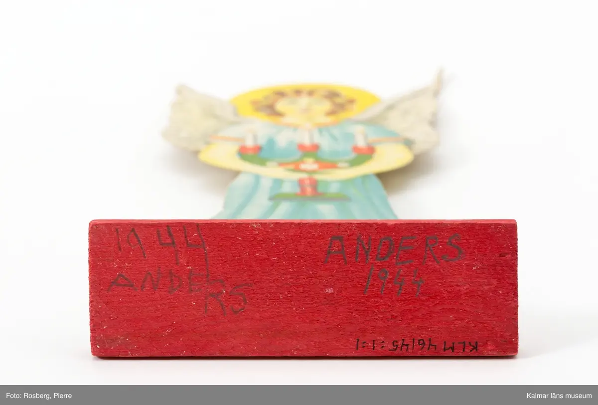 KLM 46145:1:1-2 Ängel av papper, på fot av trä. Två stycken änglar, tryckta på kraftig papp, med framsida på båda sidorna. Ängeln håller en trearmad ljusstake framför sig med båda händerna. Foten rödmålad. Signerade längst ner till vänster: Jenny Nyström. :1:1 På träfotens undersida text, Anders 1944, två gånger. Datering, senast 1944.