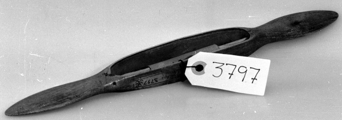 Avlang vevskyttel med flate håndtak i hver ende. Det er skåret ut et hull på midten med spile som det er surret et papir rundt.