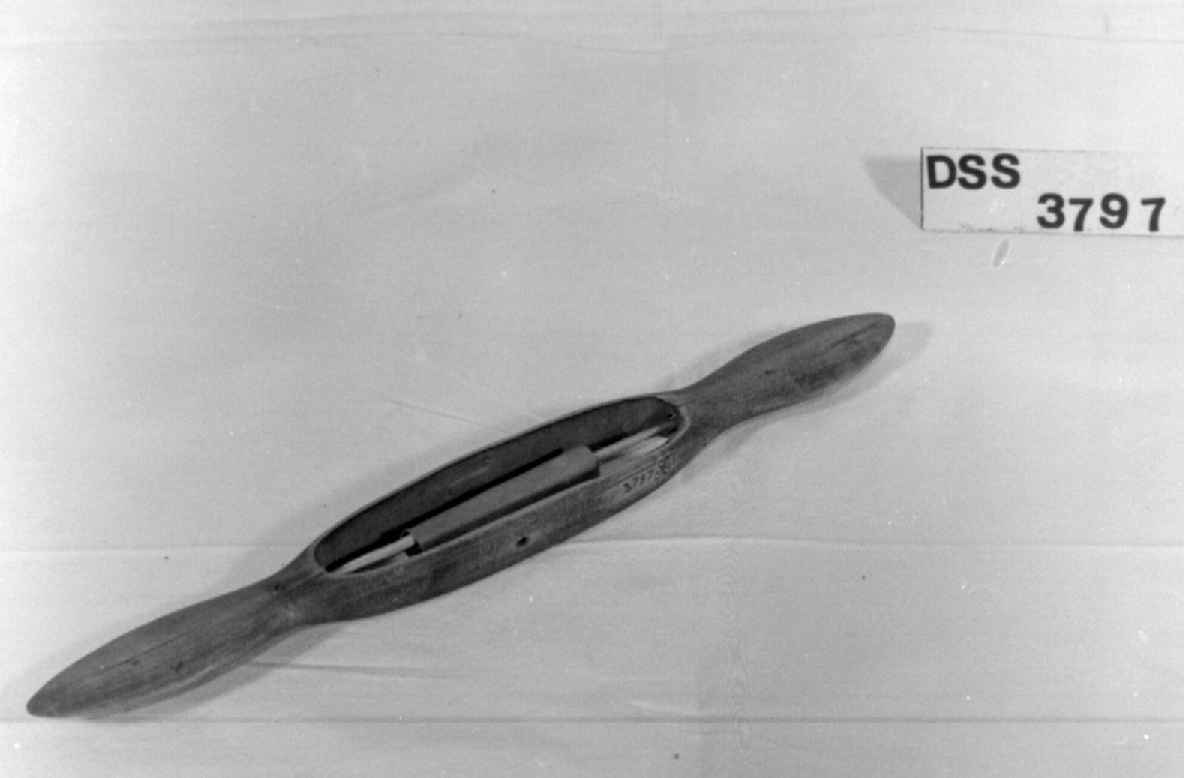 Avlang vevskyttel med flate håndtak i hver ende. Det er skåret ut et hull på midten med spile som det er surret et papir rundt.