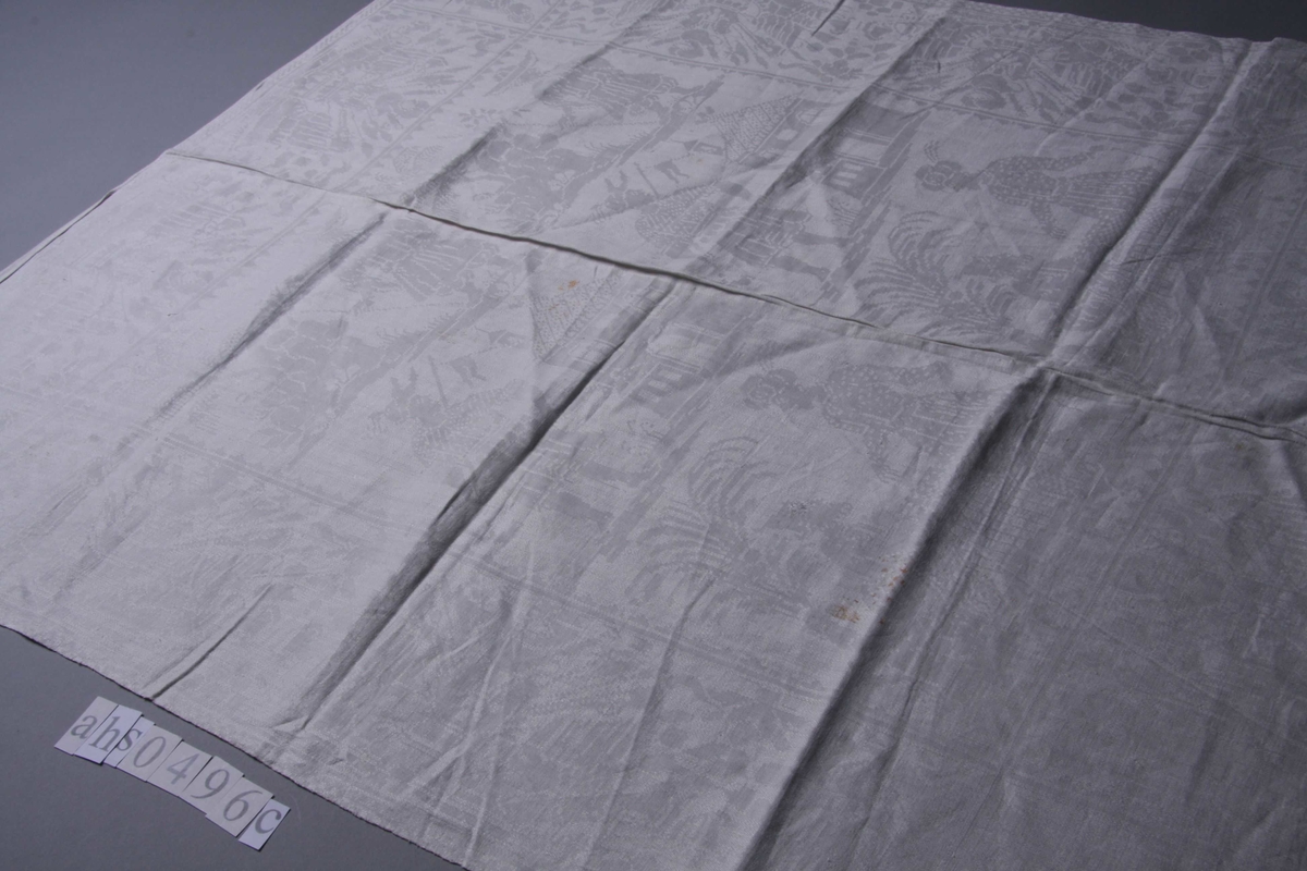 Hvit lindamask med sjakkbrett-bord langs alle fire sider. Og bred bord innenfor med trofèoppstillinger. I hvert hjørne en figur med allongeparykk. På midtpartiet en sittende og en ståendefigur ved siden, og øverst stående figur med stor hatt og tregruppe. Mønster fra beg. av 1700. Serviettene har smal fall på kortsidene.
