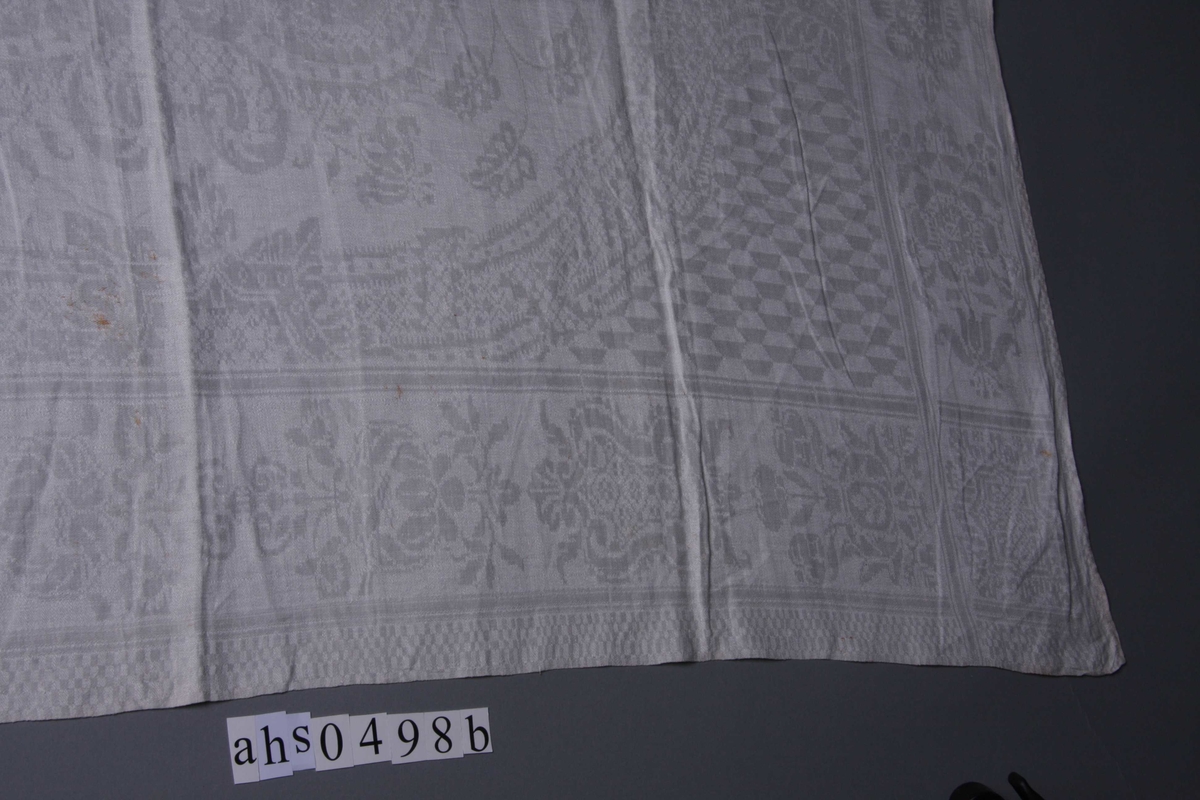 Hvit lindamask med sjakkbrett-bord på langsidene. Innenfor en bred stilisert blomsterbord med enkelte skjold-liknende motiver. Midtpartiet består av et stort dekorativt våpenskjold i ramme av stiliserte blomster og geometriske figurer. Hjørnene er utfylt med rutemønster av trapes-formete figurer. Servietten har smal fall på kortsidene og er merket.