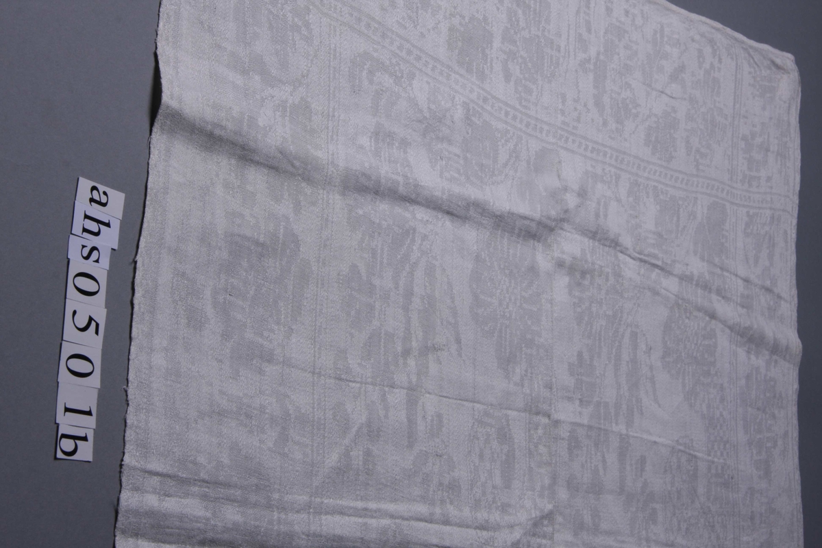 Hvit lindamask med stilisert blomsterbord langs kanten mellom to smalere stripeborder. Smal fall på kortsidene og monogram: BM, speilvendt.