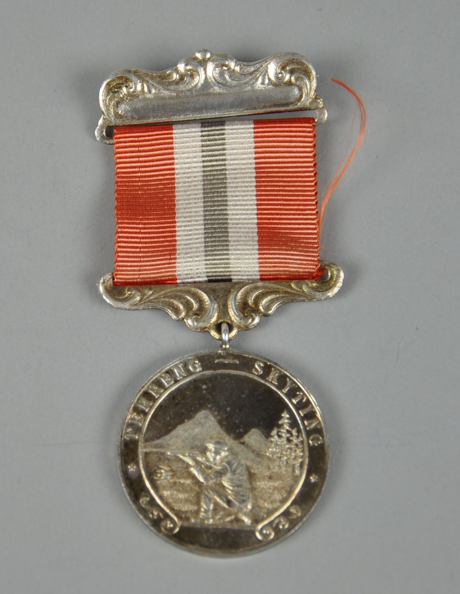 Medalje med skiskytter som sitter på kne og sikter med geværet, skog og fjellmotiv i bakgrunnen. Nål med bånd i rødt, hvit og blått (blå farge er falmet).