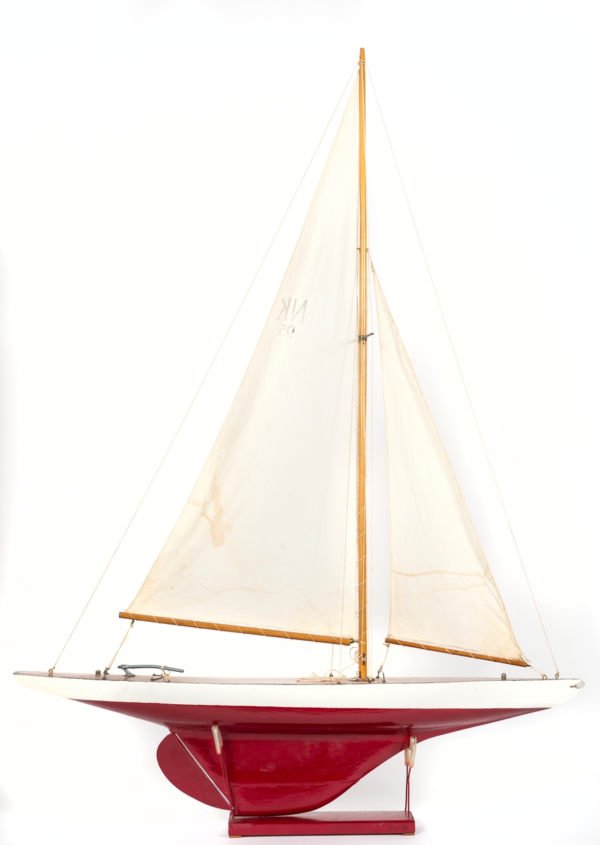 Modell av segelbåt, leksak. Tillverkad av NK senast 1935. 
Vitt skrov med röd bottenmålning. Fernissat nåtat däck och blyköl.