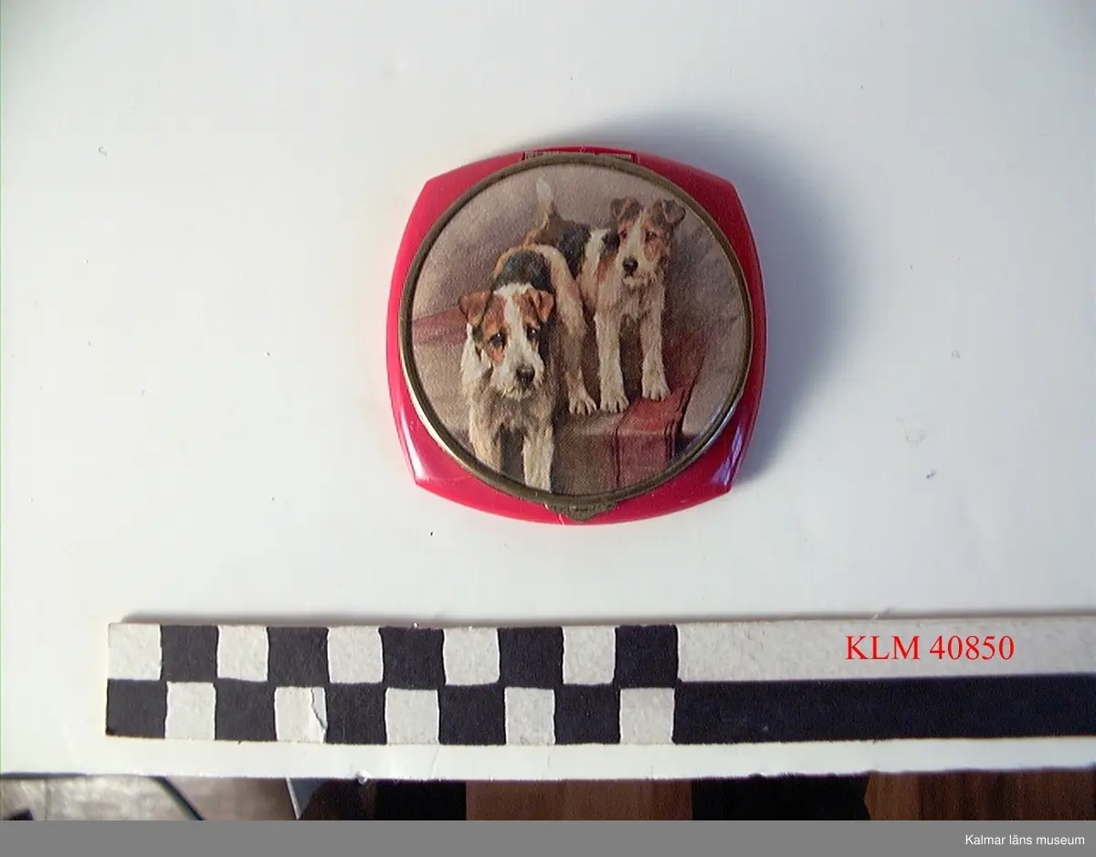 KLM 40850 Puderdosa, plast, glas. Kvadratisk med något rundade sidor, röd plast. Runt lock med spegel på insidan. På lockets utsida bild av två hundar, foxterrier.