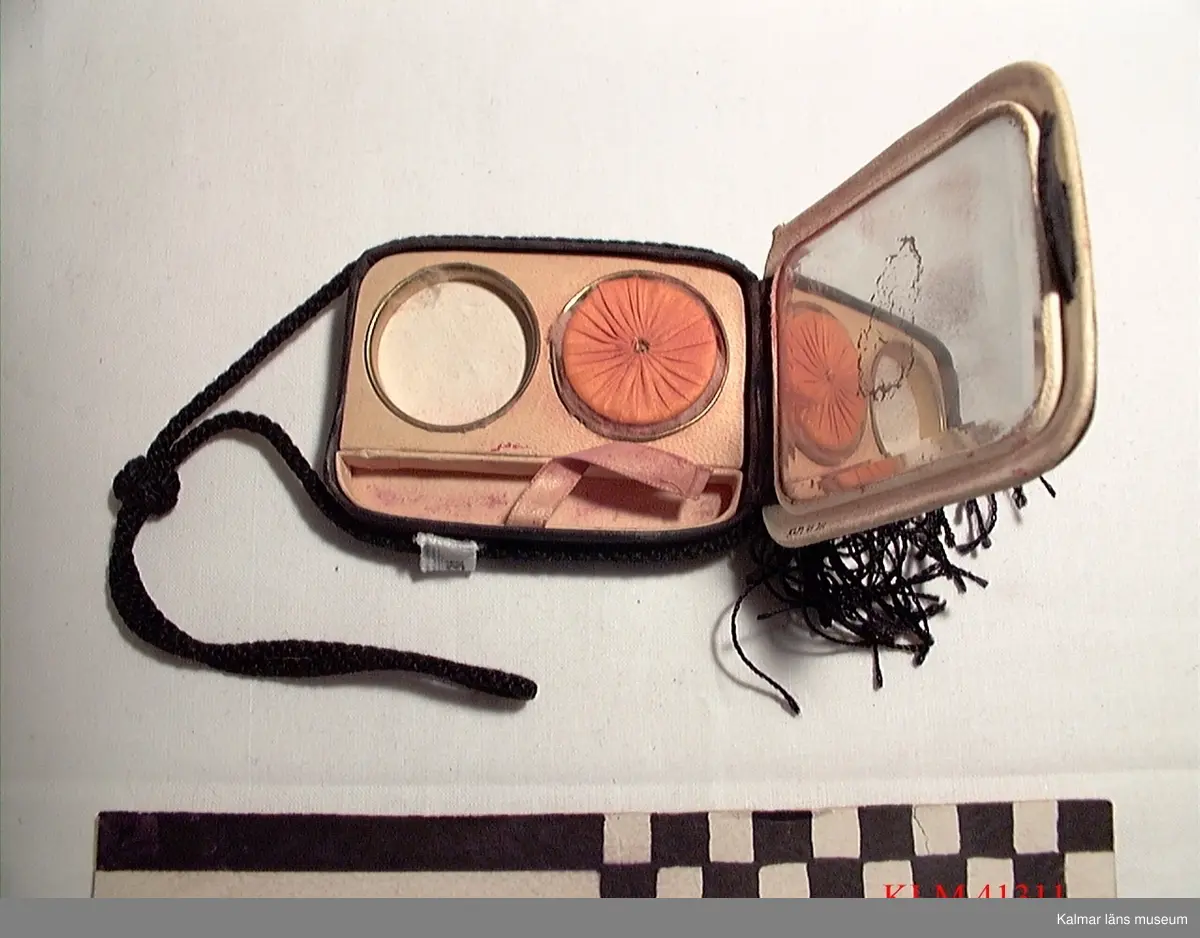 KLM 41311 Puderdosa, textil, spegelglas. Rektangulär, klädd med svart tyg, kantad med svart snodd, tofs. Inuti två runda fördjupningar med vitt respektive rött puder/rouge, rund vippa av tyg, spegel i locket.