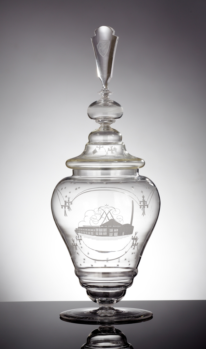Pokal med lock.
Pokalen är balusterformad med en frikostig diameter på dess övre del.
Pokalens undre del smalnar av kraftigt och avslutas med en relativt stor fot.
Locket presenteras i olika etager och avslutas med en hög knopp.
På pokalen graverade girlanger och stjärnor, samt ett monogram och en bild på Elme glasbruk.
På locket graverad girlang och stjärnor, och på knoppen graverat "Elme".