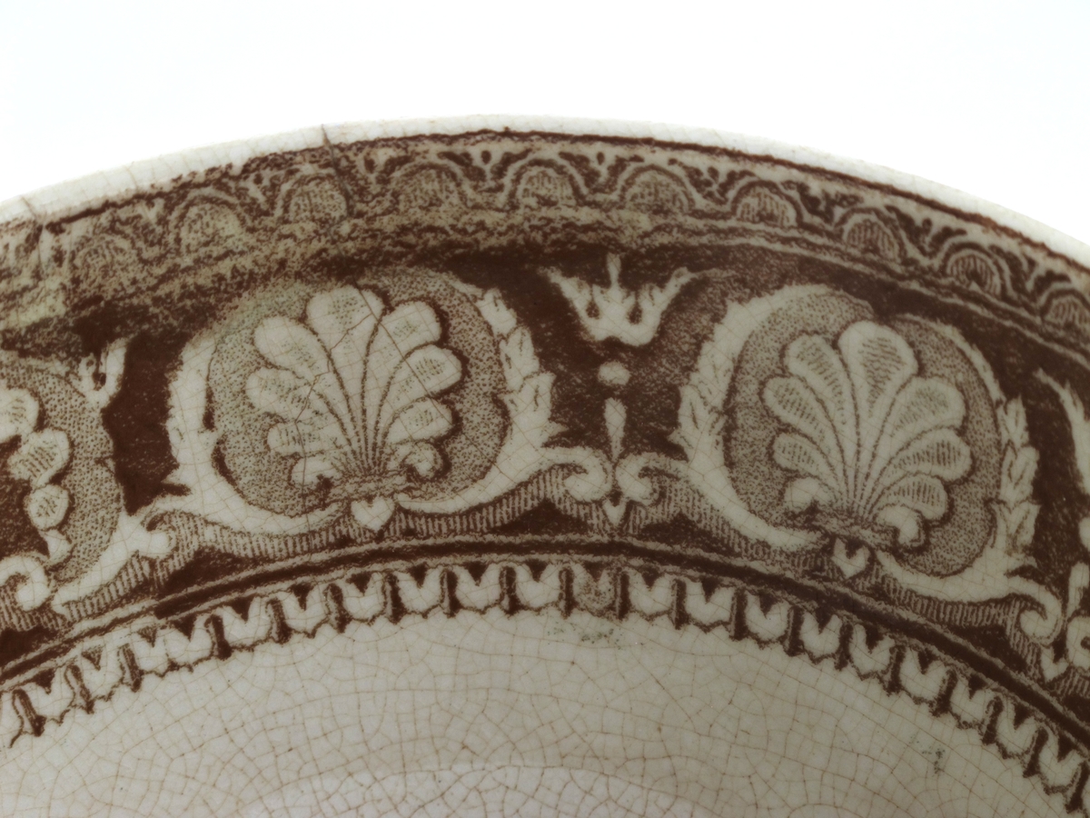 Bolle, stentøy, Buede sider, innsvunget fotring; trykt mønster i  brunt: Innv. og utv. bred ornamental bord m. skjell og "Garrya Eliptica" i bunnen lignende dekor, her er skjellene plassert i korsform i et rundt felt. Tilst. juli 1961: Krakellert.