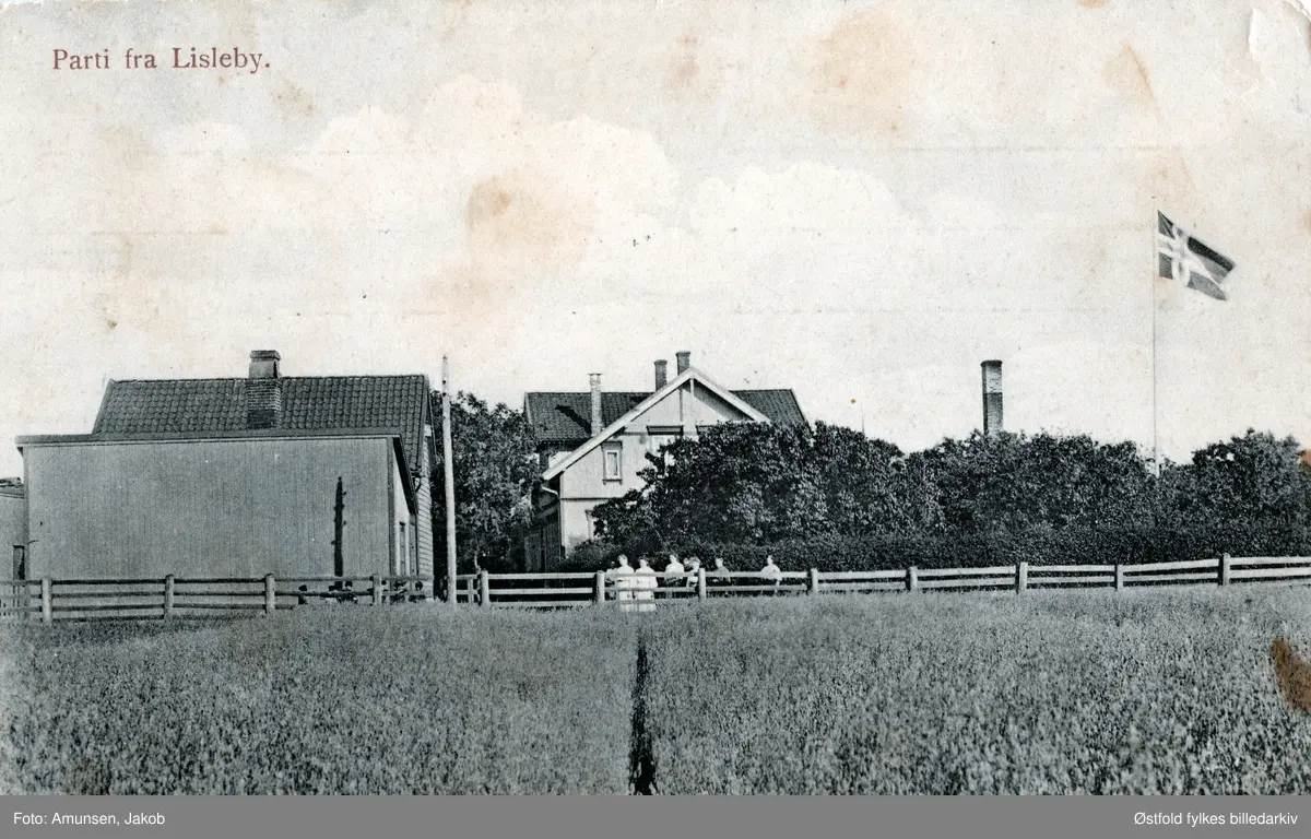 Postkort med parti fra Lisleby.
Postkort med motiv fra Lisleby, landhandel, og bakeri, ca. 1905-10 mot Selbak fergested. Aasta Gjøby arbdeidet der. Bygningen brant engang før 1920.
I dag ligger NorBetong Ferdigbetong på stedet, dagens adresse er Dr. Opsandsvei 33.
Gruppe med 8 personer står langs gjerdet.

Huset var et av husene som gikk med i brannen på bordtomten til Lisleby bruket 10 mai 1915. Der alle bordhusene og 11 beboelseshus gikk med i brannen. Jacob Amundsen tok etter det over Fredrikstad Fargehandel i Baggaden 2, som Farmanns gate het den gangen. (Johreid, info 2018).