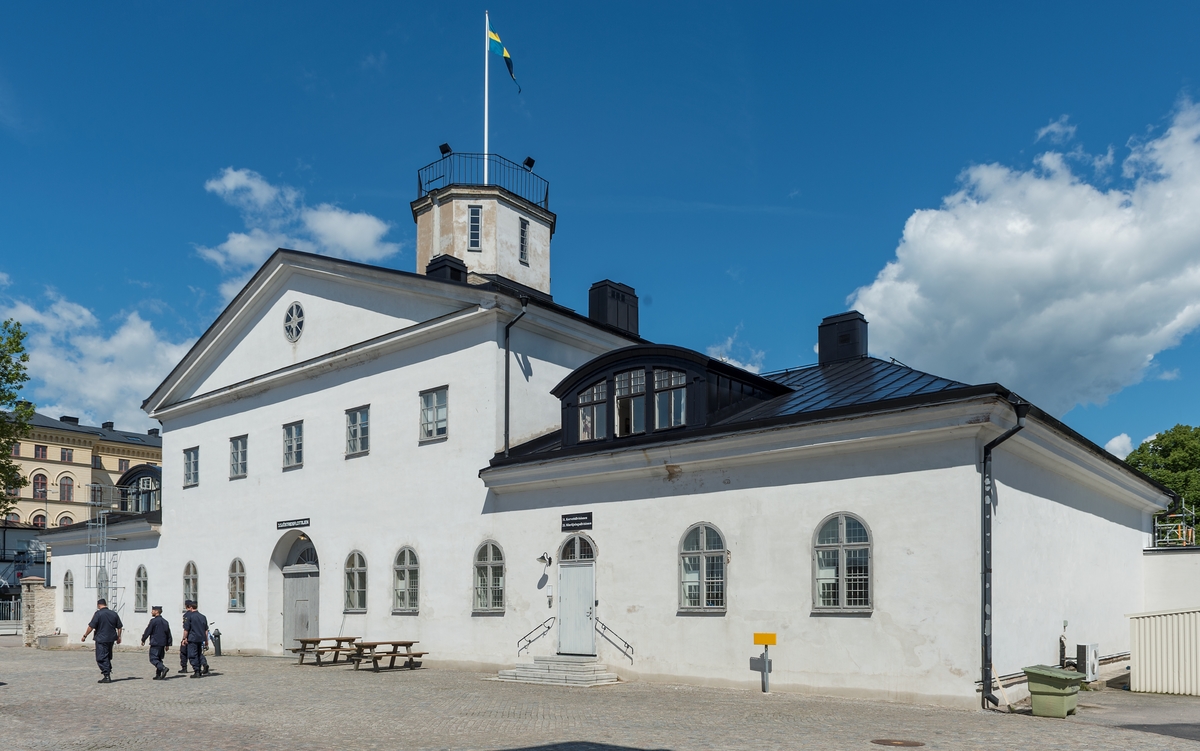 Envåningsbyggnad i grå puts och plåttak. Mittparti i två våningar med en arkadgång av fem bågar vid huvudentrén.
