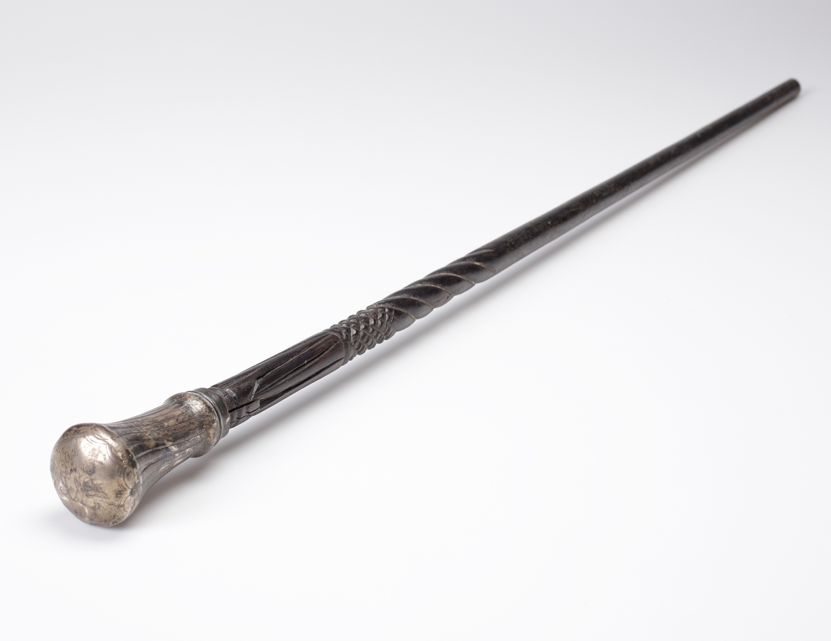 Promenadkäpp, s.k. spanskrör. 
Mörk, fernissad (?) käpp med delvis spiralvridet, snidat mönster och diamantskärningar. Knopp av silver med randad, något svängd dekor. På knoppens ovansida graverat ägarmonogram: "SEL" (?). Längst ner på käppen en skoning av ben. Stämplar längs nedre kanten av knoppen.
