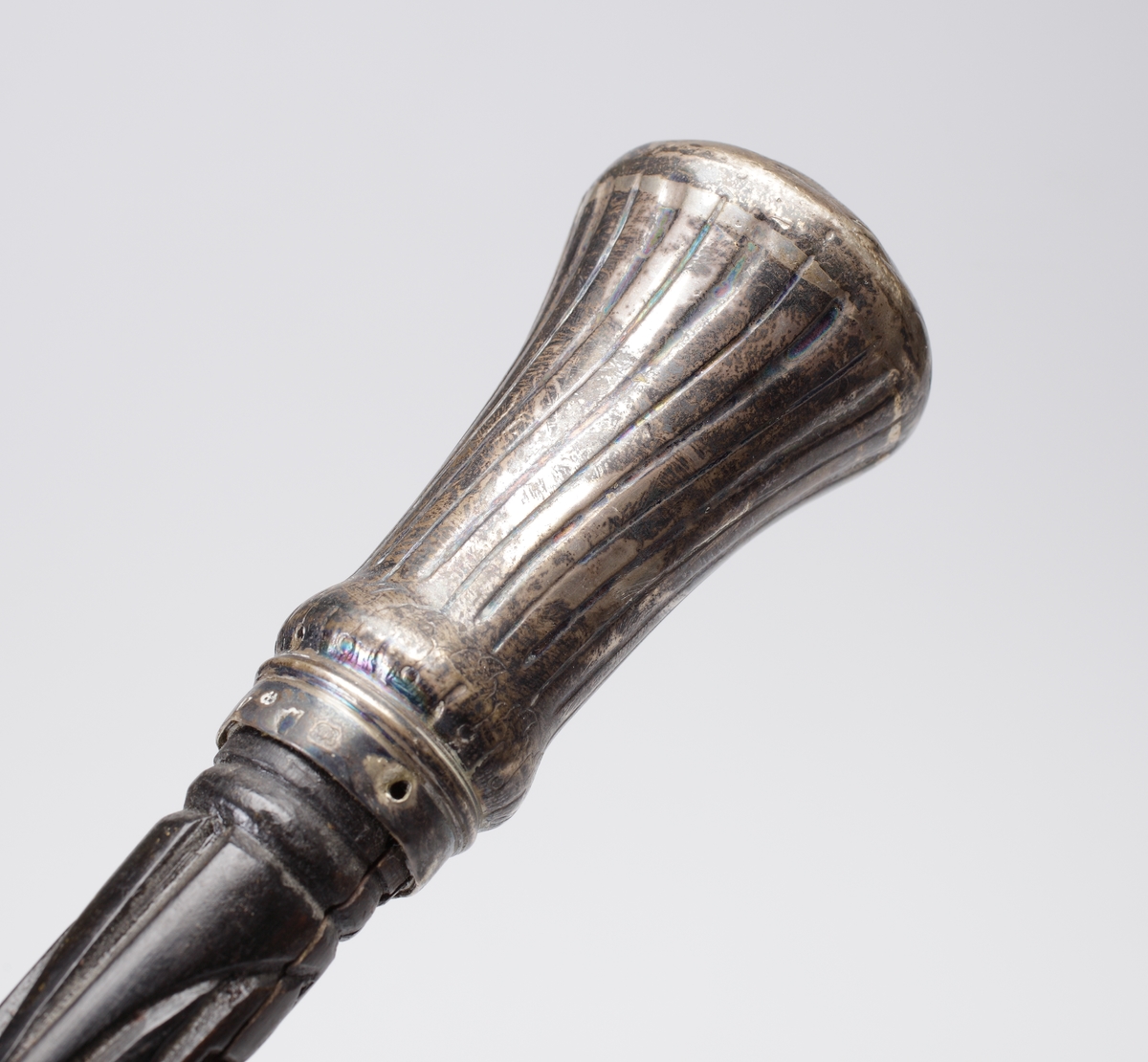 Promenadkäpp, s.k. spanskrör. 
Mörk, fernissad (?) käpp med delvis spiralvridet, snidat mönster och diamantskärningar. Knopp av silver med randad, något svängd dekor. På knoppens ovansida graverat ägarmonogram: "SEL" (?). Längst ner på käppen en skoning av ben. Stämplar längs nedre kanten av knoppen.