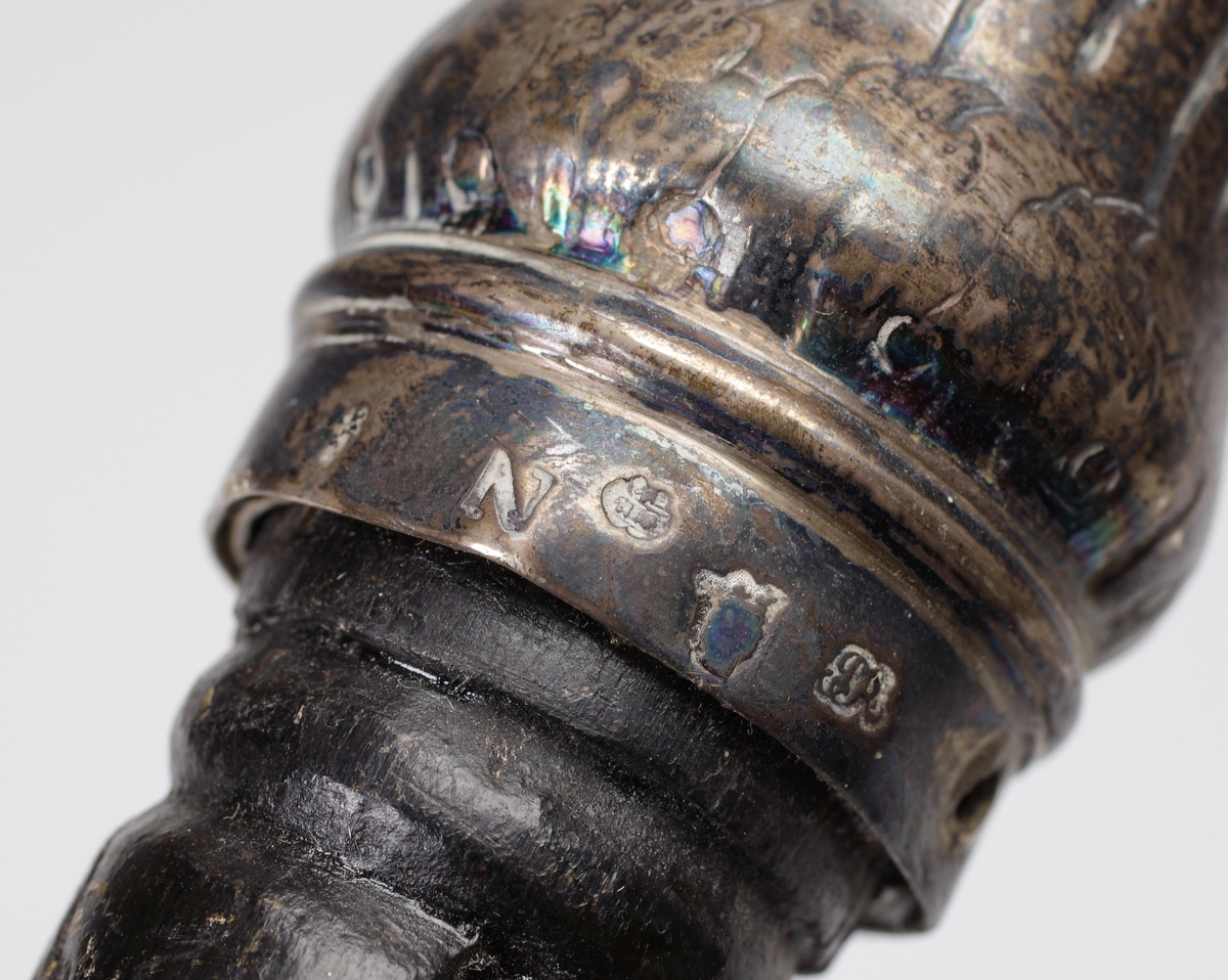 Promenadkäpp, s.k. spanskrör. 
Mörk, fernissad (?) käpp med delvis spiralvridet, snidat mönster och diamantskärningar. Knopp av silver med randad, något svängd dekor. På knoppens ovansida graverat ägarmonogram: "SEL" (?). Längst ner på käppen en skoning av ben. Stämplar längs nedre kanten av knoppen.