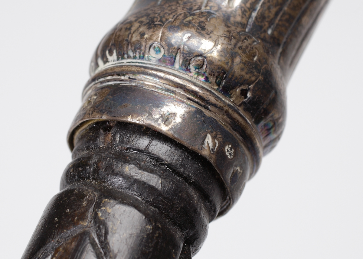 Promenadkäpp, s.k. spanskrör. 
Mörk, fernissad (?) käpp med delvis spiralvridet, snidat mönster och diamantskärningar. Knopp av silver med randad, något svängd dekor. På knoppens ovansida graverat ägarmonogram: "SEL" (?). Längst ner på käppen en skoning av ben. Stämplar längs nedre kanten av knoppen.