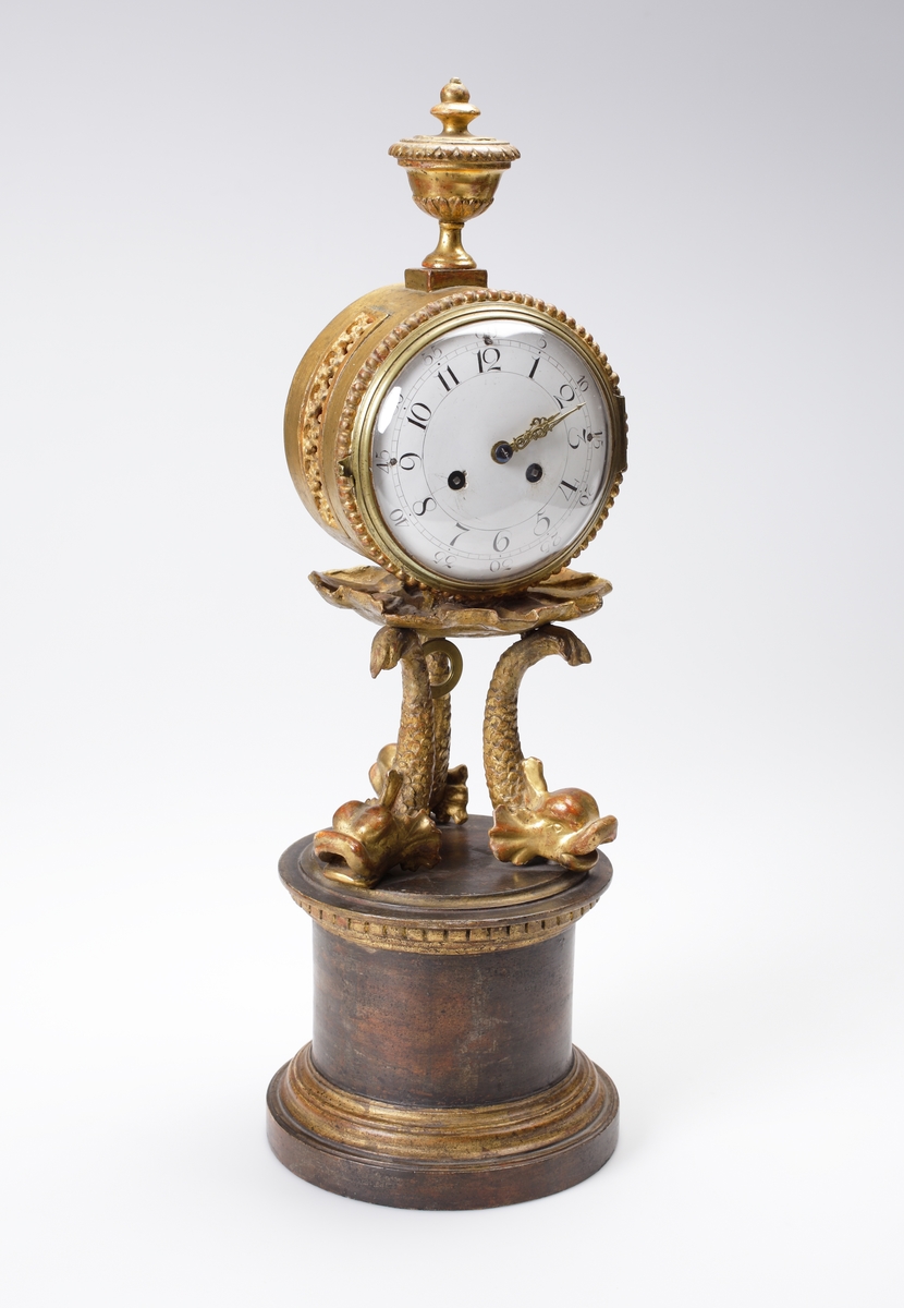 Bordspendyl / Bordsstudsare.
Vit emaljerad urtavla försedd med arabiska siffror i en rund, guldbronserad överdel av trä, som vilar på tre snidade delfiner. Högst upp en guldbronserad urna. 
Cylindrisk fot av trä, målad i rödbrun marmorimitation med guldbronserade partier. Tillhörande urnyckel av järn.