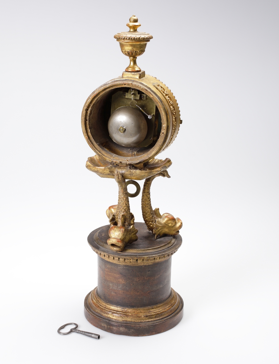 Bordspendyl / Bordsstudsare.
Vit emaljerad urtavla försedd med arabiska siffror i en rund, guldbronserad överdel av trä, som vilar på tre snidade delfiner. Högst upp en guldbronserad urna. 
Cylindrisk fot av trä, målad i rödbrun marmorimitation med guldbronserade partier. Tillhörande urnyckel av järn.