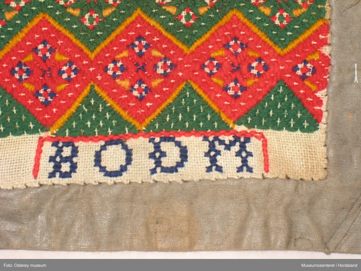 Brodert med ullgarn i plattsaum. Alt er handlaga. Hovudmotiv er små åttebladroser i border. Oppe er det påsydd eit blått silkeband med eit breitt gullband (i forgylt sølvtråd) utanpå silkebandet. Alt er montert på eit tynt, vokset bomullslerret. Oppe på baksida er det eit ekstra bomullstoff (blåstripet). Bokstavane BODM er broderte inn nede på bringeduken i kross-sting.