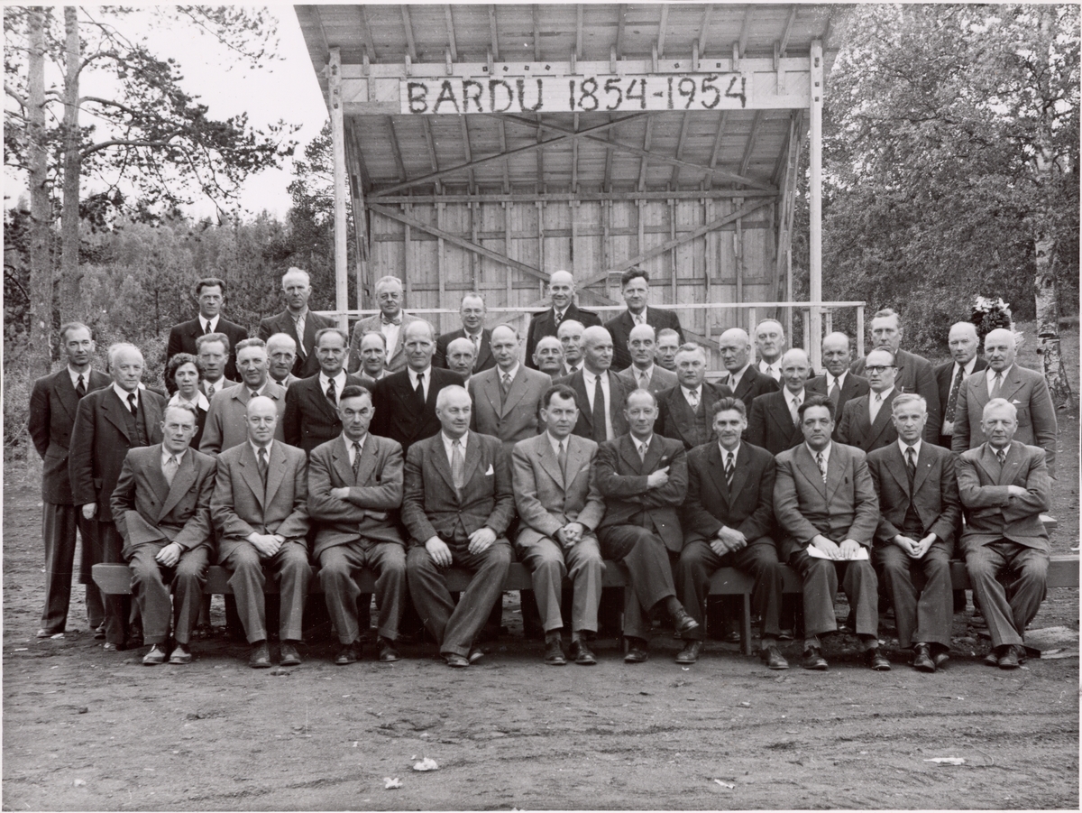 Gruppebilde tatt foran en paviljong dekorert med teksten"Bardu 1854-1954".