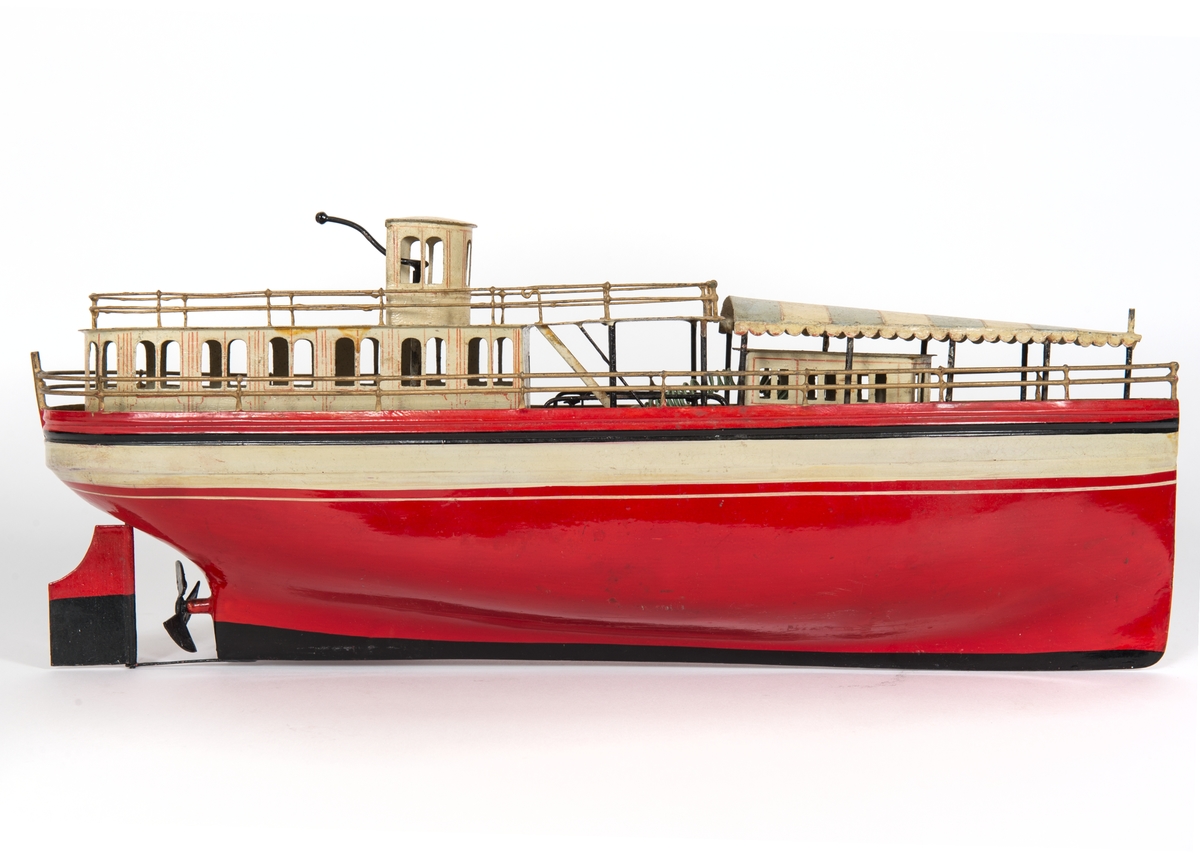 Fartygsmodell. Leksaksbåt av plåt. Rödmålat skrov med svartvitt bälte över vattenlinjen. Rak förstäv, häckakter, roder och en trebladig propeller. Förligt däckshus överspänt med "suntält" av plåt, akterut passagerarsalong, navigationshytt. Propellermaskineriet består av en tvåpolig elektrisk motor driven av ett De Lanche element. Gråmålat skrå.