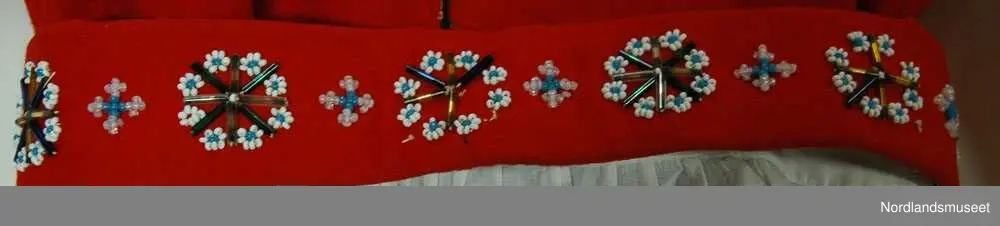 Perle broderi forran på brystet og på belte. På brystet er perlene formet som et hjerte og det er perler inni og på siden. Motivet på skjorten og forkleet er et hardanger motiv som er meget fint. På luen, vesten og skjørtet er det en gull bård som går rundt hele kanten.