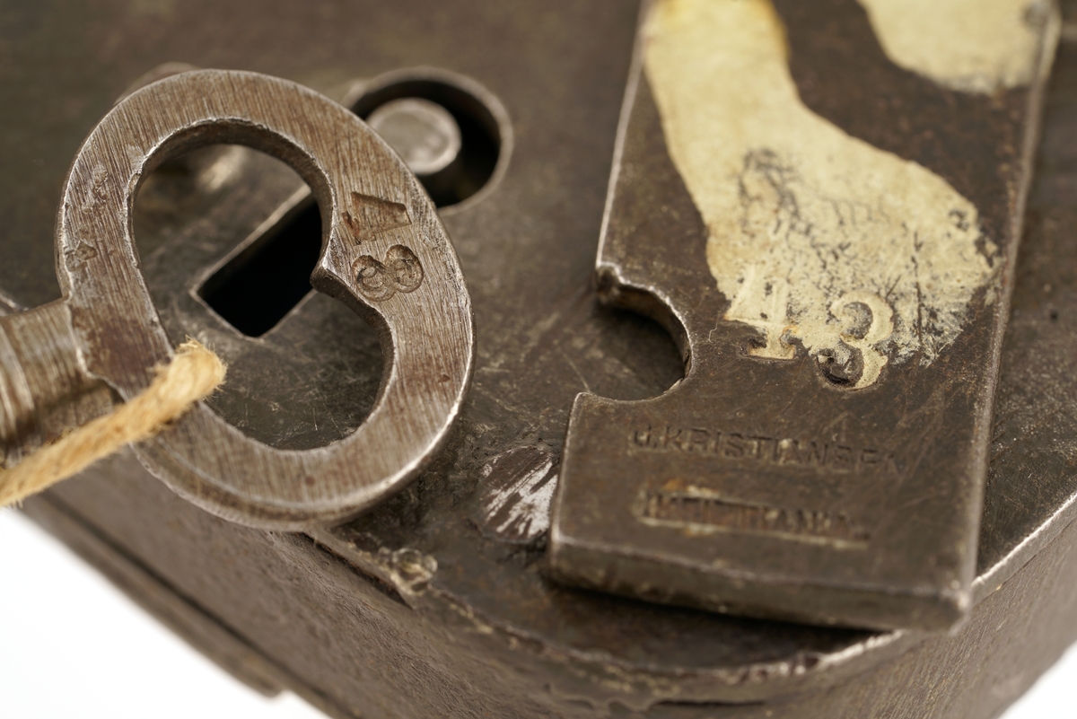 Jern, firkantet, svingbart lokk over nøkkelhullet.
Nøkkel, jern, firkantet skjegg, gjennomboret.
Papirlapp knyttet fast i nøkkel med snor.