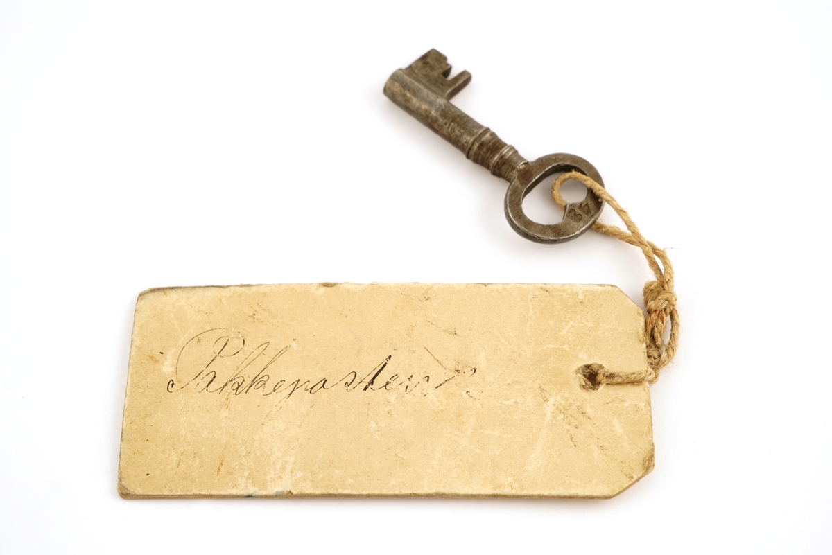 Jern, firkantet, svingbart lokk over nøkkelhullet.
Nøkkel, jern, firkantet skjegg, gjennomboret.
Papirlapp knyttet fast i nøkkel med snor.