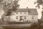 Mangårdsbygganden till Herrestad mellangård. Enligt påskrift togs bilden 1879. Vid tiden ägdes gården av Nils Gustaf Eriksson och dennes maka Josefina Andersdotter. Möjligtvis är det gårdsägaren som sitter i kostym och hatt på bilden. Övriga personer på bilden stämmer överens med familjens sammansättning vid tiden; hustrun, barnen Anna Josefina f. 1859, Emma Desideria f. 1861, Alida Charlotta f. 1863, Erik Gustaf f. 1869  och Helena Elisabeth f. 1870.