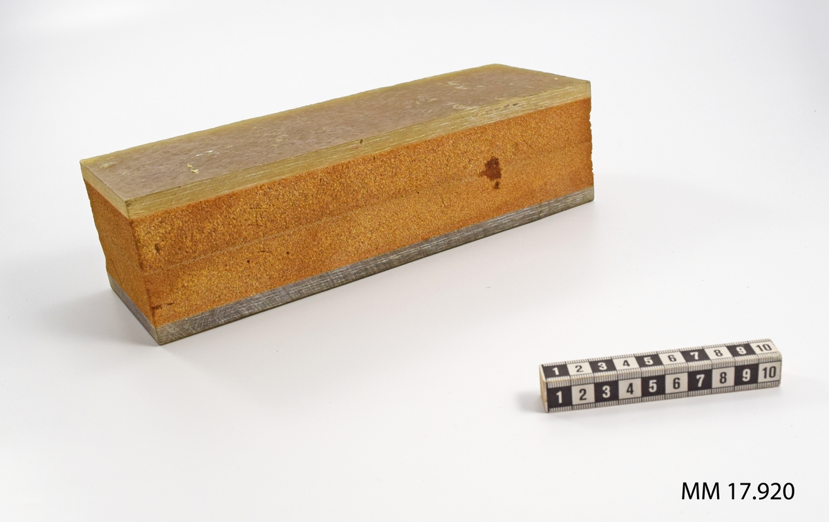 Gjutet laminatstycke, rektangulärt utsågat, bestående av kompakta ytskikt, Tj = 10 mm, som utgör bordläggningen. Mellan dessa ett poröst skikt, kärnan, sammansatt av två skikt. Brun färg.