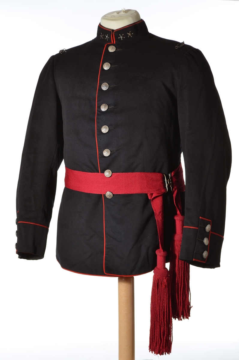 Enkeltspent jakke til uniform i mørk blå ull, med rød kanting og sølvnapper. Åtte knapper i fronten. Kraven er opprettstående og med to sølvstjerner på hver side. Skuldrene har tre påsydde hemper og metallkjede. Knappene er dekorert med norsk løve. Baksiden har splitt og tre par knapper. Rød kantdekor. Nederst på ermene er det tre smale bånd med knapp og røde kantbånd. Innvendig er jakken helforet med rødt stoff og lyst stripet stoff i ermene. Opphengshemper i ermene. Fronten er vattert. Innvendig hempe i midjen. To hekter i halsen for lukning av krave. Lomme i foret på hver side av splitten.

Bukse i mørk blå ull med to lommer foran og en bak. Gylf lukkes med metallhekte og fire plastknapper. Spenne øverst mot linning bak. Seks knapper i linning. En mangler. Striper av rødt stoff i sidene. Fôr i linning og litt nedenfor linning.

Belte i burgunder ull med store dusker i hver ende og messingspenne. Beltet festes rundt livet over jakken.