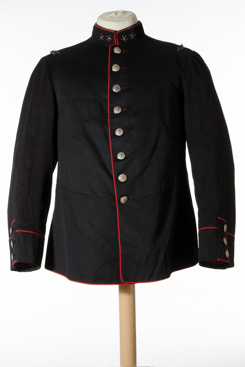 Enkeltspent jakke til uniform i mørk blå ull, med rød kanting og sølvnapper. Åtte knapper i fronten. Kraven er opprettstående og med to sølvstjerner på hver side. Skuldrene har tre påsydde hemper og metallkjede. Knappene er dekorert med norsk løve. Baksiden har splitt og tre par knapper. Rød kantdekor. Nederst på ermene er det tre smale bånd med knapp og røde kantbånd. Innvendig er jakken helforet med rødt stoff og lyst stripet stoff i ermene. Opphengshemper i ermene. Fronten er vattert. Innvendig hempe i midjen. To hekter i halsen for lukning av krave. Lomme i foret på hver side av splitten.

Bukse i mørk blå ull med to lommer foran og en bak. Gylf lukkes med metallhekte og fire plastknapper. Spenne øverst mot linning bak. Seks knapper i linning. En mangler. Striper av rødt stoff i sidene. Fôr i linning og litt nedenfor linning.

Belte i burgunder ull med store dusker i hver ende og messingspenne. Beltet festes rundt livet over jakken.